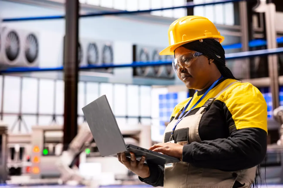 Worker wearing a yellow hard hat and uniform uses a laptop inside an industrial factory setting