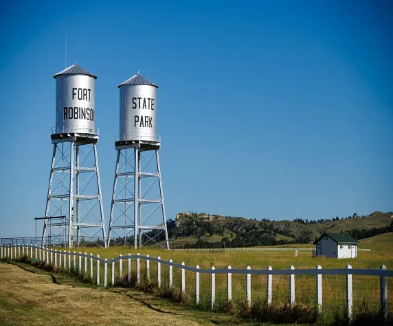 Two silver water towers labeled Fort Robinson and State Park stand in a grassy field with white fences and a small building under a clear blue sky.
