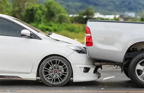 White sedan car rear-ended silver pickup truck on a road with greenery and hills in the background